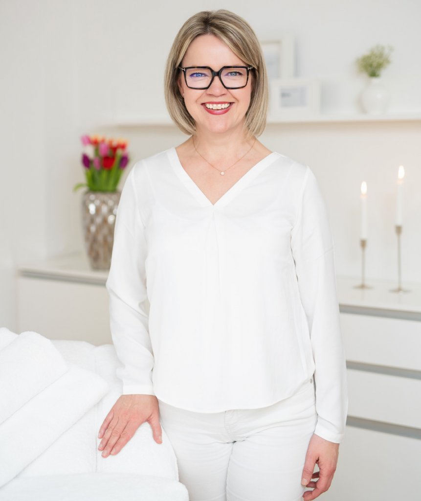 Die lächelnde Katrin Streit mit hellblonden Haaren und einer Brille, die in einem weißen Raum steht. Sie trägt eine weiße Bluse mit V-Ausschnitt und weiße Hosen. Im Hintergrund sind ein Strauß bunter Tulpen, weiße Kerzen und eine weiße Wand zu sehen.